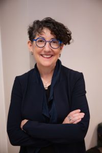 The late gender-smart investing pioneer Suzanne Biegel wears bright blue round glasses, with short curly dark hair. She is smilling with arms folded